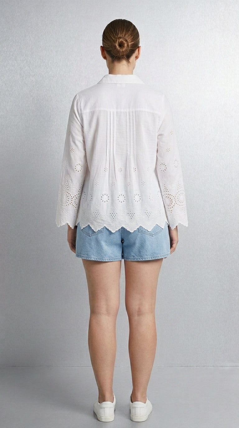 A woman viewed from the back wearing a white long-sleeve eyelet blouse with a scalloped hem and light blue denim shorts against a light gray studio background.