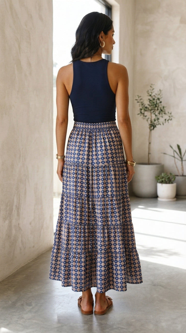 A woman is shown from behind wearing a sleeveless navy blue top and a long, pleated skirt with a blue, white, and brown geometric pattern. She is standing in a bright, modern room with textured beige walls and potted plants.