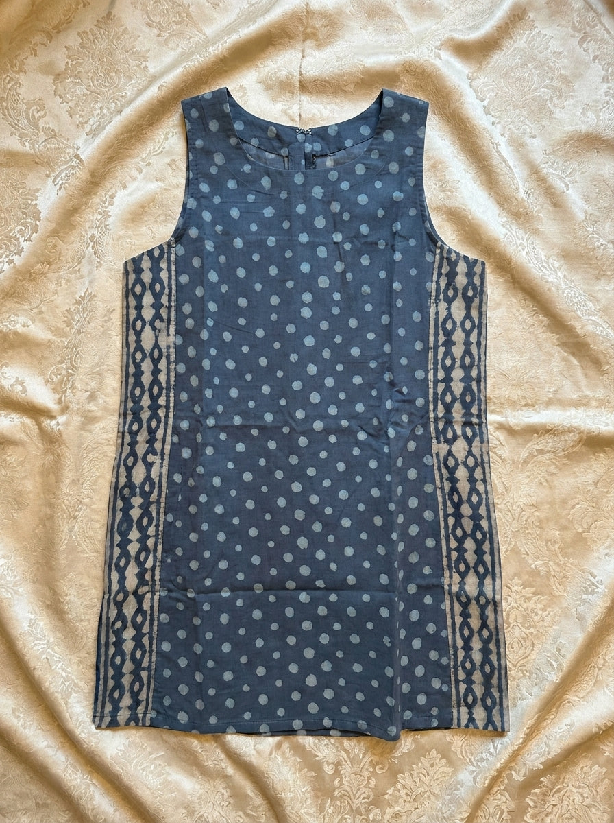 A sleeveless blue dress with a white polka dot pattern and geometric side panels, laid flat on a beige textured fabric background.