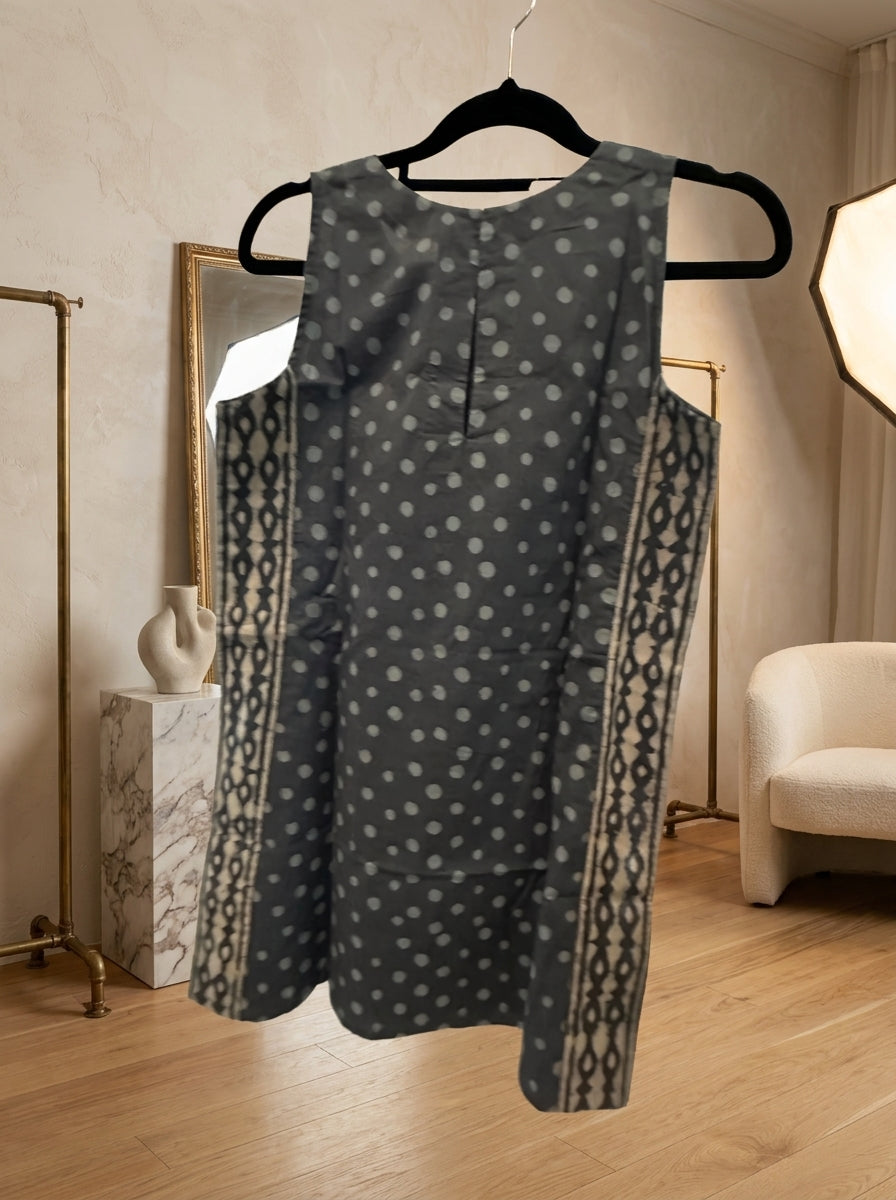 A sleeveless dark gray dress with white polka dots and patterned side panels, hanging on a black hanger in a styled indoor room with wood floors and neutral decor.