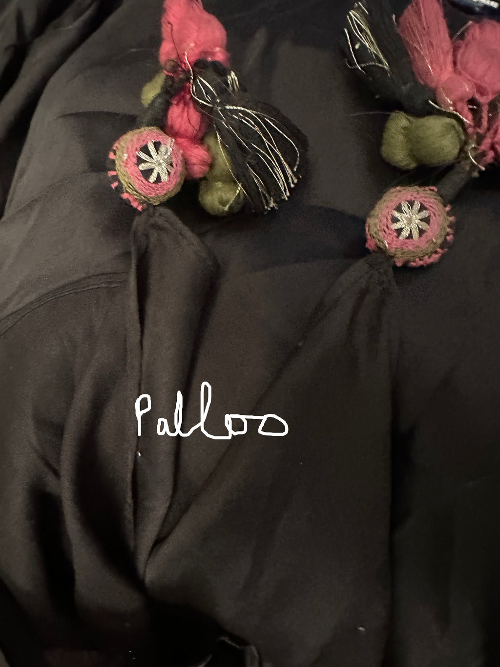 A close-up of a black garment featuring decorative tassel and pom-pom embellishments in pink, black, and olive green.