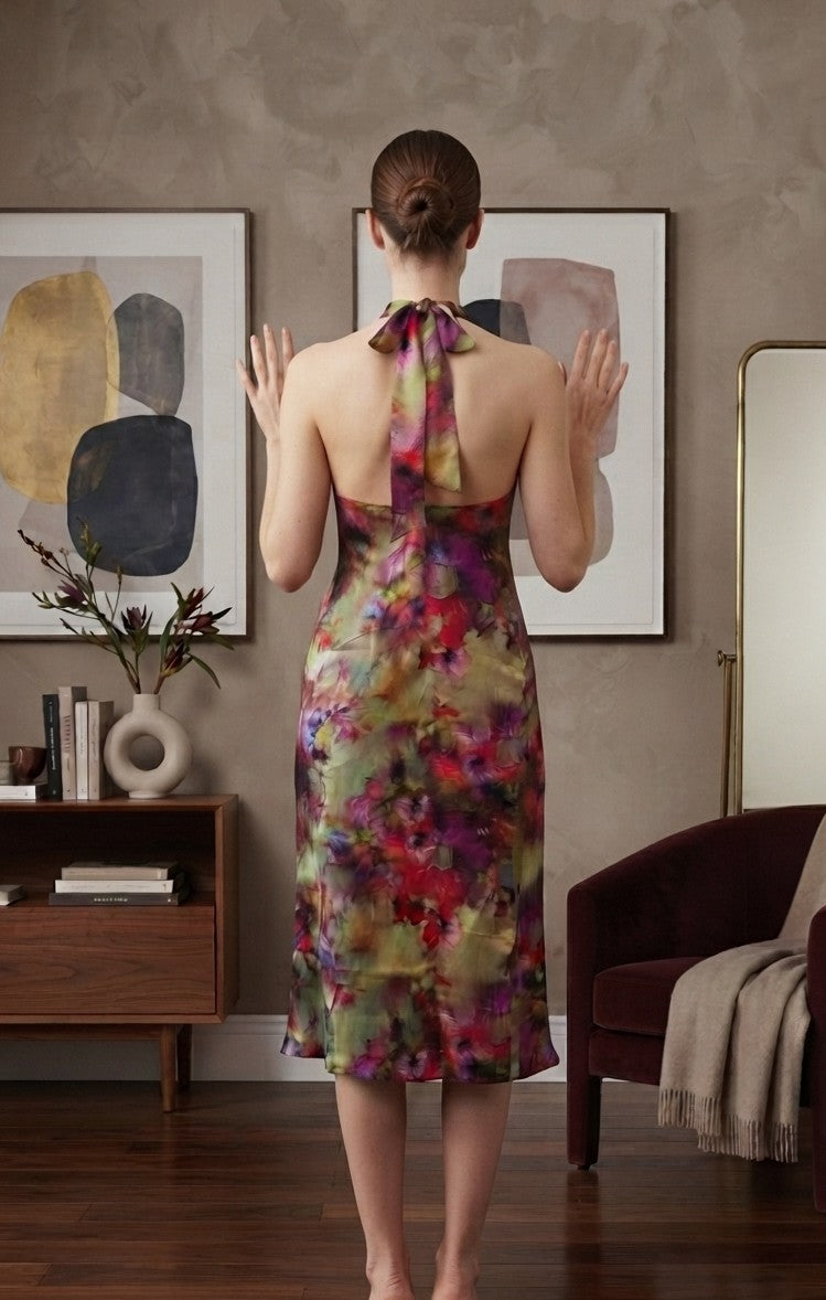 A woman stands with her back to the camera wearing a sleeveless, backless midi dress with a colorful floral print and a tie-neck detail, set in a styled living room interior.