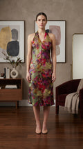 A woman stands in a furnished room wearing a sleeveless halter-neck midi dress with a multicolor floral print in red, pink, purple, green, and gold tones.