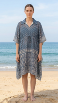 A woman stands on a sandy beach wearing a loose, short-sleeve kaftan-style dress with a blue and white pattern. The ocean and sky are visible in the background.