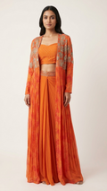 A woman modeling a traditional Indian outfit consisting of an orange crop top, matching pleated skirt, and a sheer orange dupatta with gold embroidery, set against a plain light beige background.