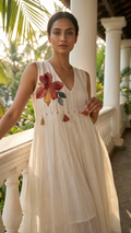 A woman wearing a sleeveless cream-colored dress with a large red and orange floral embroidery on the chest, standing on a balcony with white pillars and green foliage in the background.