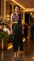 A woman models a sleeveless black evening pre draped saree with a heavily embroidered pink and gold halter crop top in a warmly lit indoor event space.