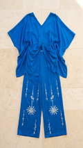 A royal blue two-piece cord set consisting of a draped short-sleeve top and matching wide-leg pants, laid flat on a beige stone tile background. The pants feature white embroidered snowflake and vertical line patterns.