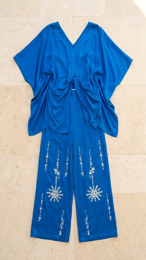 A royal blue two-piece cord set consisting of a draped short-sleeve top and matching wide-leg pants, laid flat on a beige stone tile background. The pants feature white embroidered snowflake and vertical line patterns.